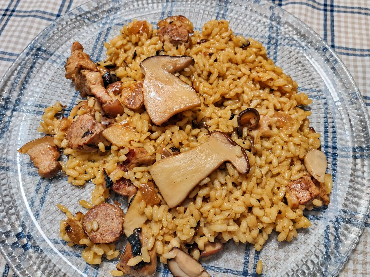 Arròs de ceps amb cansalada i rodanxes de botifarra, servit en un plat de vidre sobre unes estovalles de quadres.