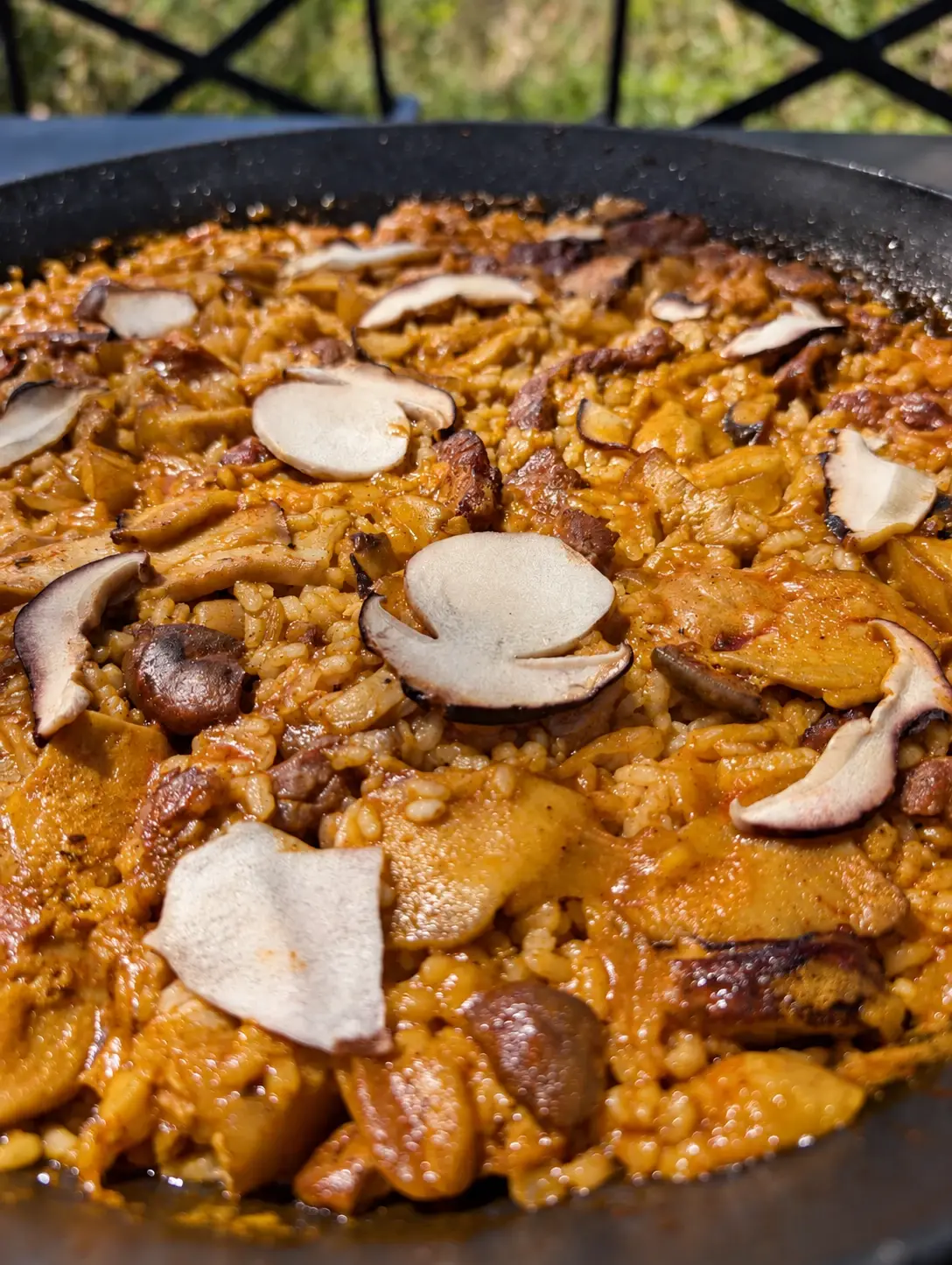 Arròs de galtes de porc amb ceps a la paella, vist de prop a l’aire lliure: arròs vermellós amb trossos de galta i de cep i làmines fines de cep per sobre.
