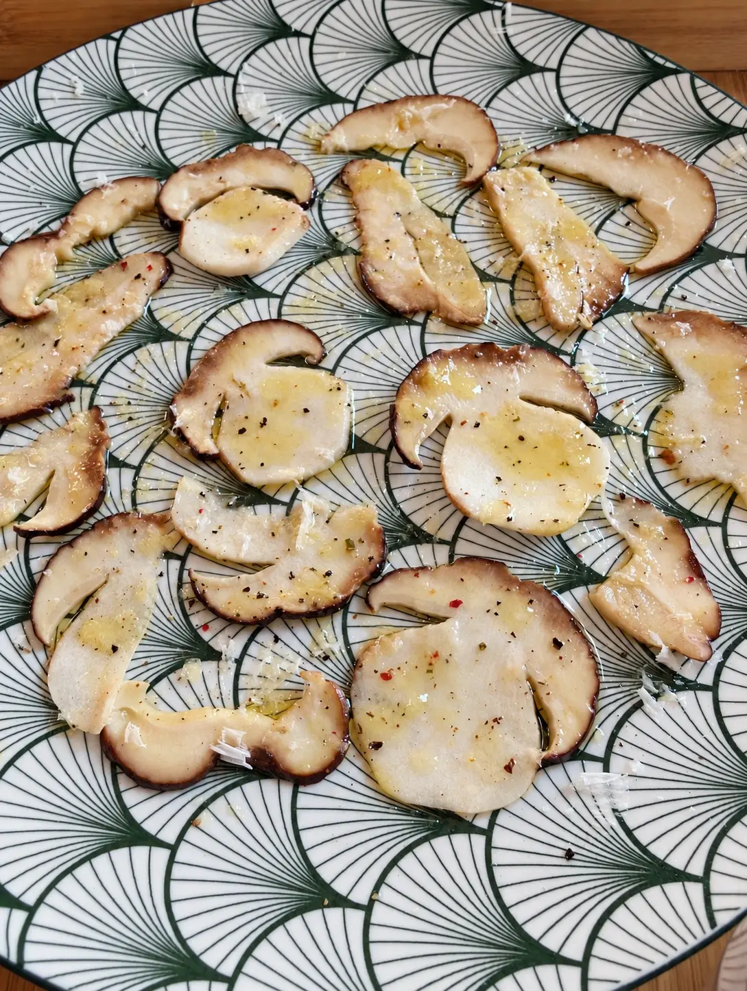 Carpaccio de ceps vist des de dalt, amb oli, pebre i formatge sobre un plat blanc amb ventalls verds.