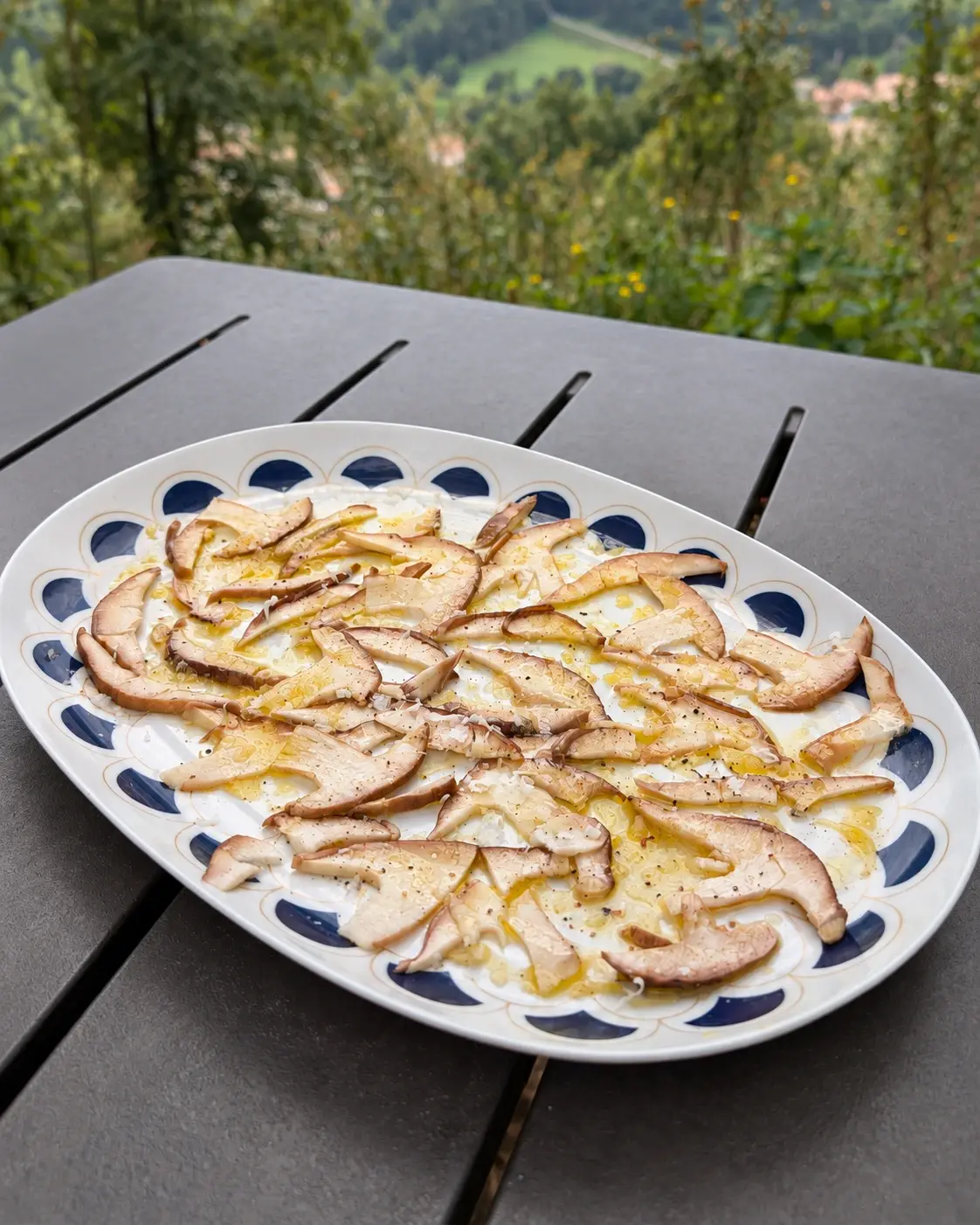 Carpaccio de ceps en una plata ovalada blanca amb la vora de mitges llunes blaves, sobre una taula exterior de llistons foscos: làmines fines de cep cru amb oli d’oliva, pebre negre i formatge ratllat.